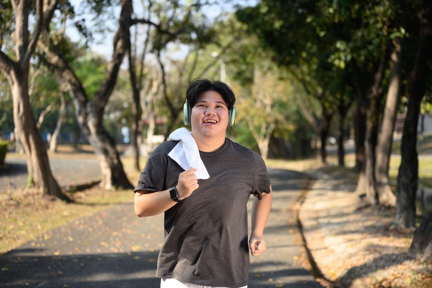 A smiling person wearing headphones and a towel around their neck jogging through a sunlit park.