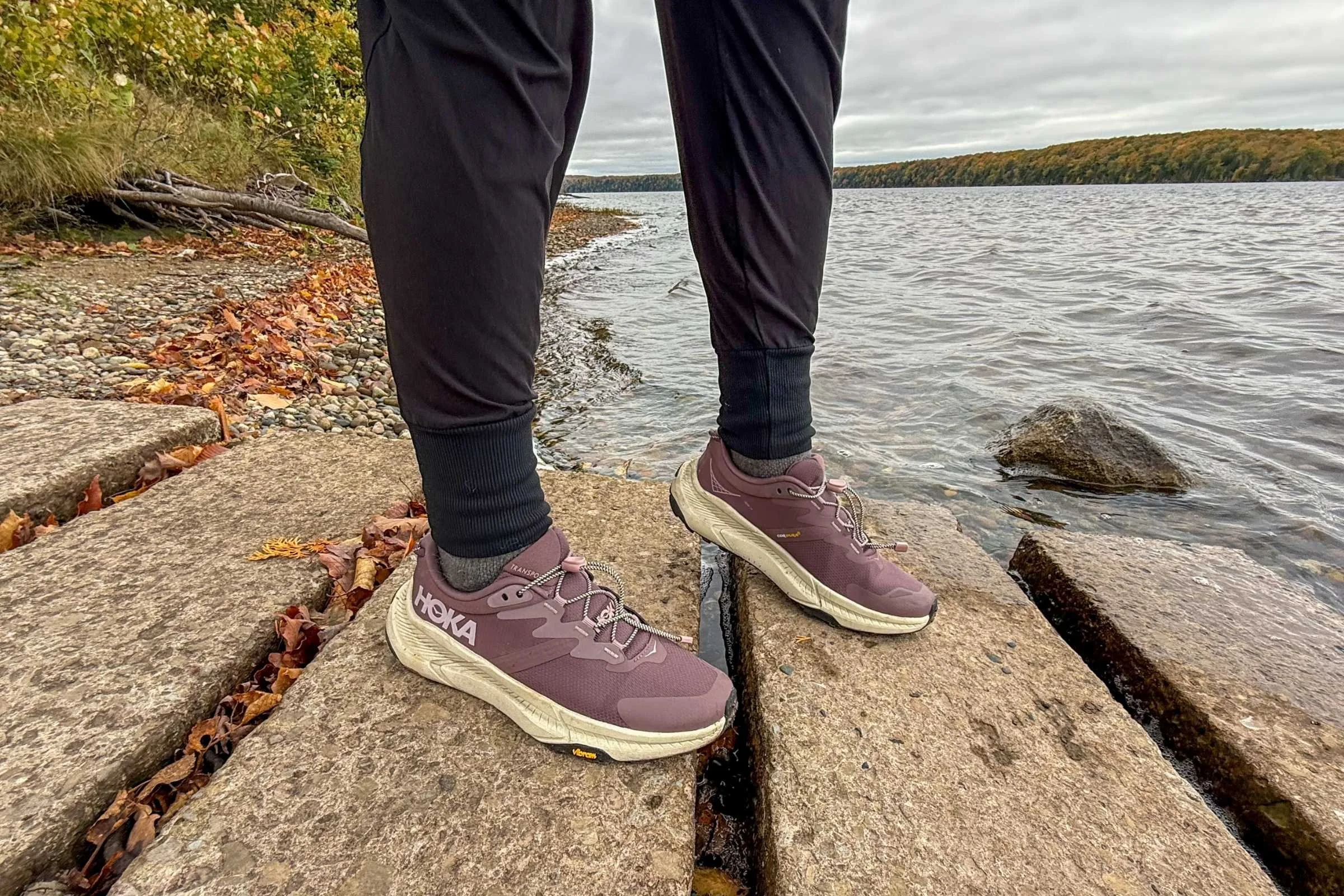 A person standing by a lake in HOKA Transport 2 shoes, suitable for mixed surfaces.