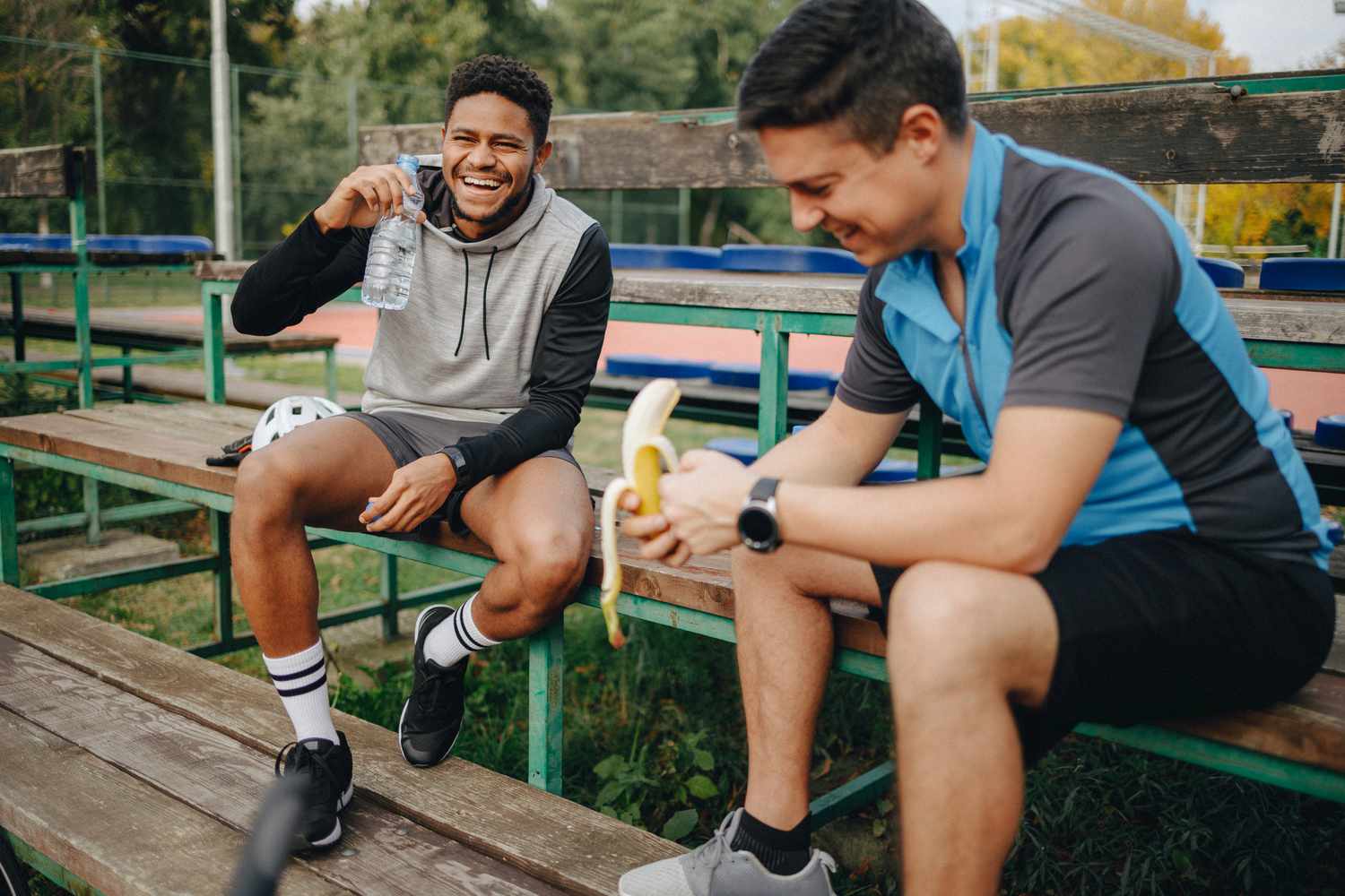 Two men in athletic wear resting after a workout with one eating a banana and the other drinking water.