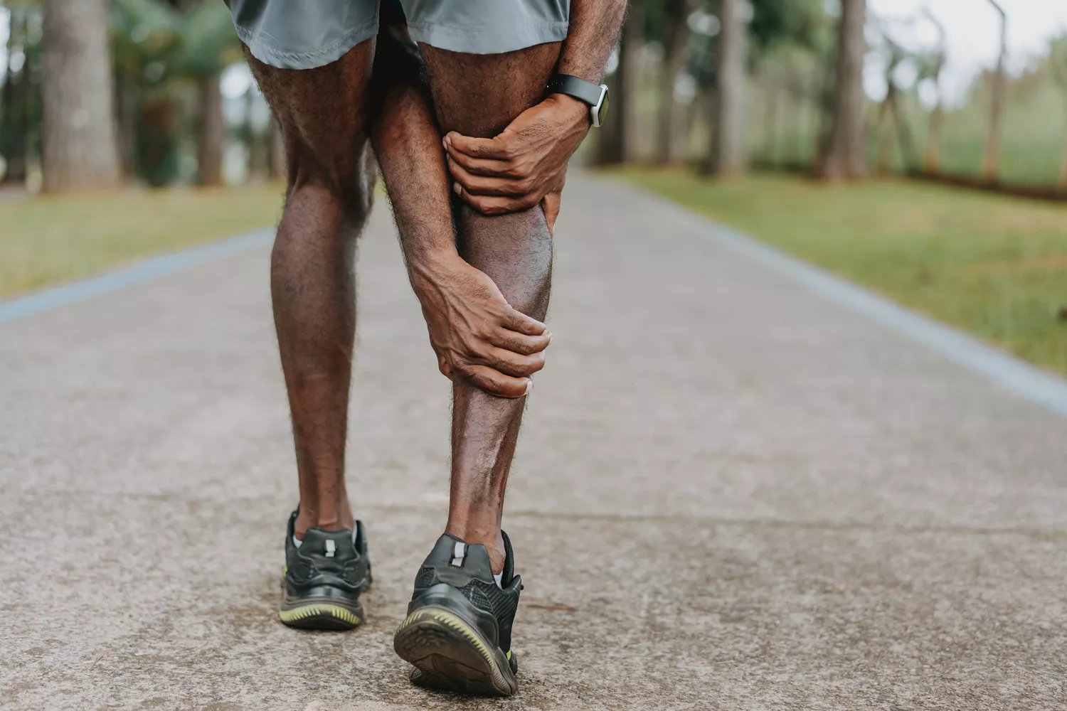A runner on an outdoor path stopping to hold their leg due to a sudden muscle spasm.