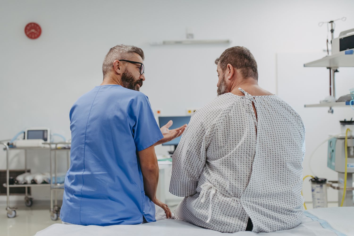 A male surgeon in blue scrubs discussing weight loss surgery options with a patient in a clinical room setting.