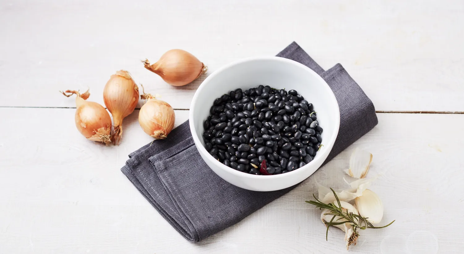 Dried black beans in a bowl surrounded by raw onions, garlic, and a sprig of rosemary.