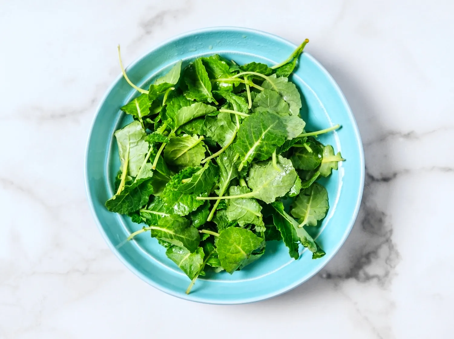 Fresh leafy greens in a white bowl on a marble surface.