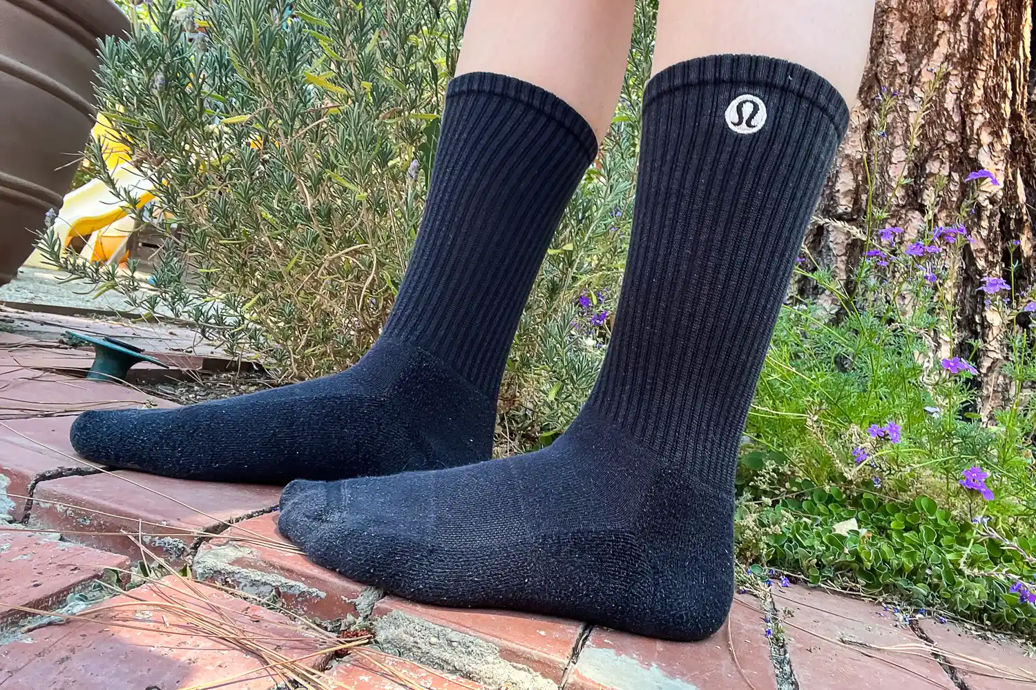 A person wearing black Lululemon crew socks standing on a brick patio in a garden.