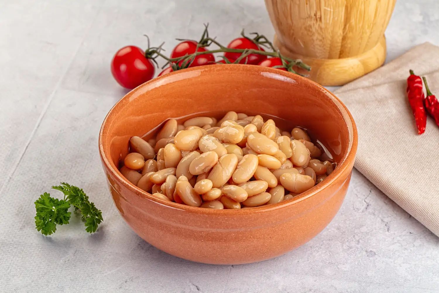White kidney beans in a bowl on a wooden table next to fresh red tomatoes and peppers.