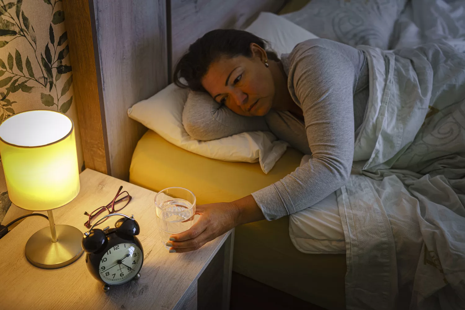 A woman reaching for a glass of water on a nightstand while in bed at night.
