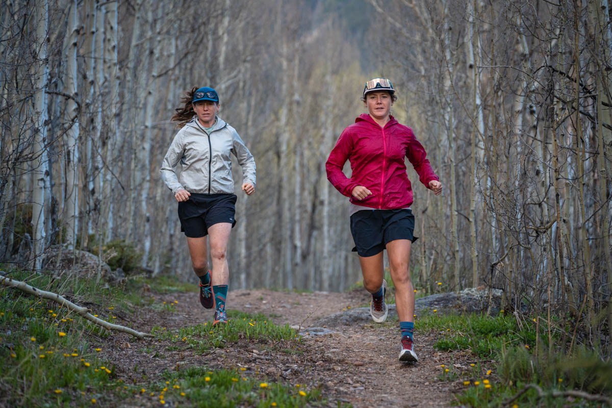 Two runners testing different technical jackets on a forested trail.