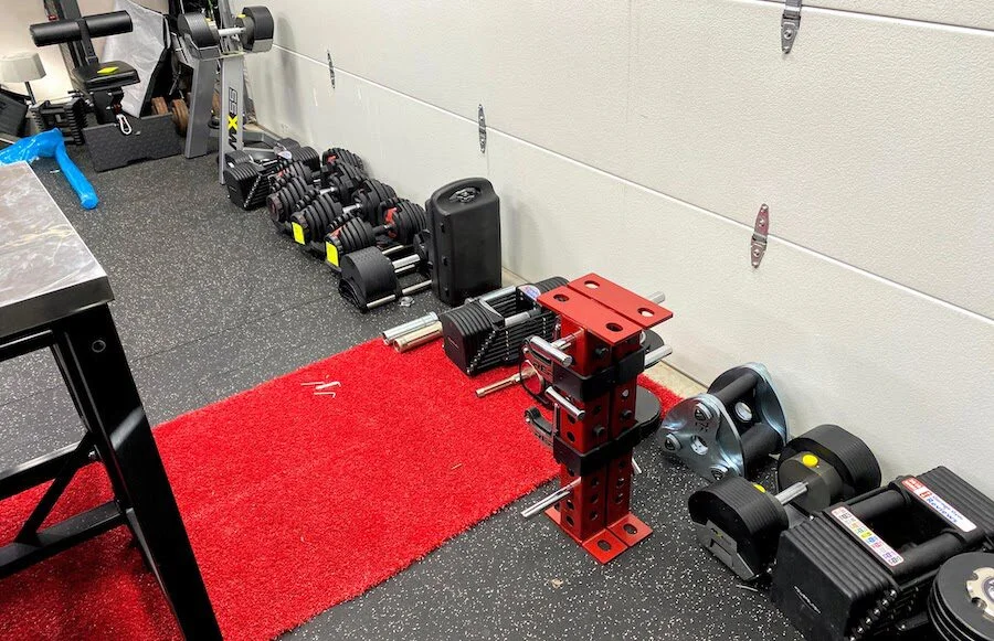 A row of various brands and models of adjustable dumbbells lined up against a wall.