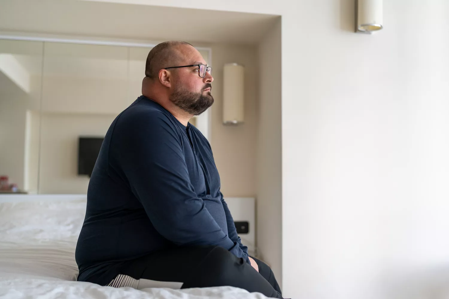 A man with a beard sitting on the edge of a bed looking contemplative and tired.