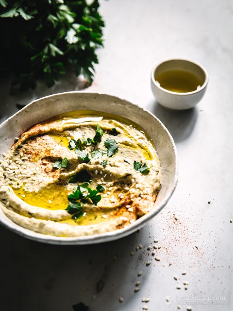 A beautifully plated bowl of zucchini hummus topped with fresh parsley and sesame seeds next to vegetable dippers.