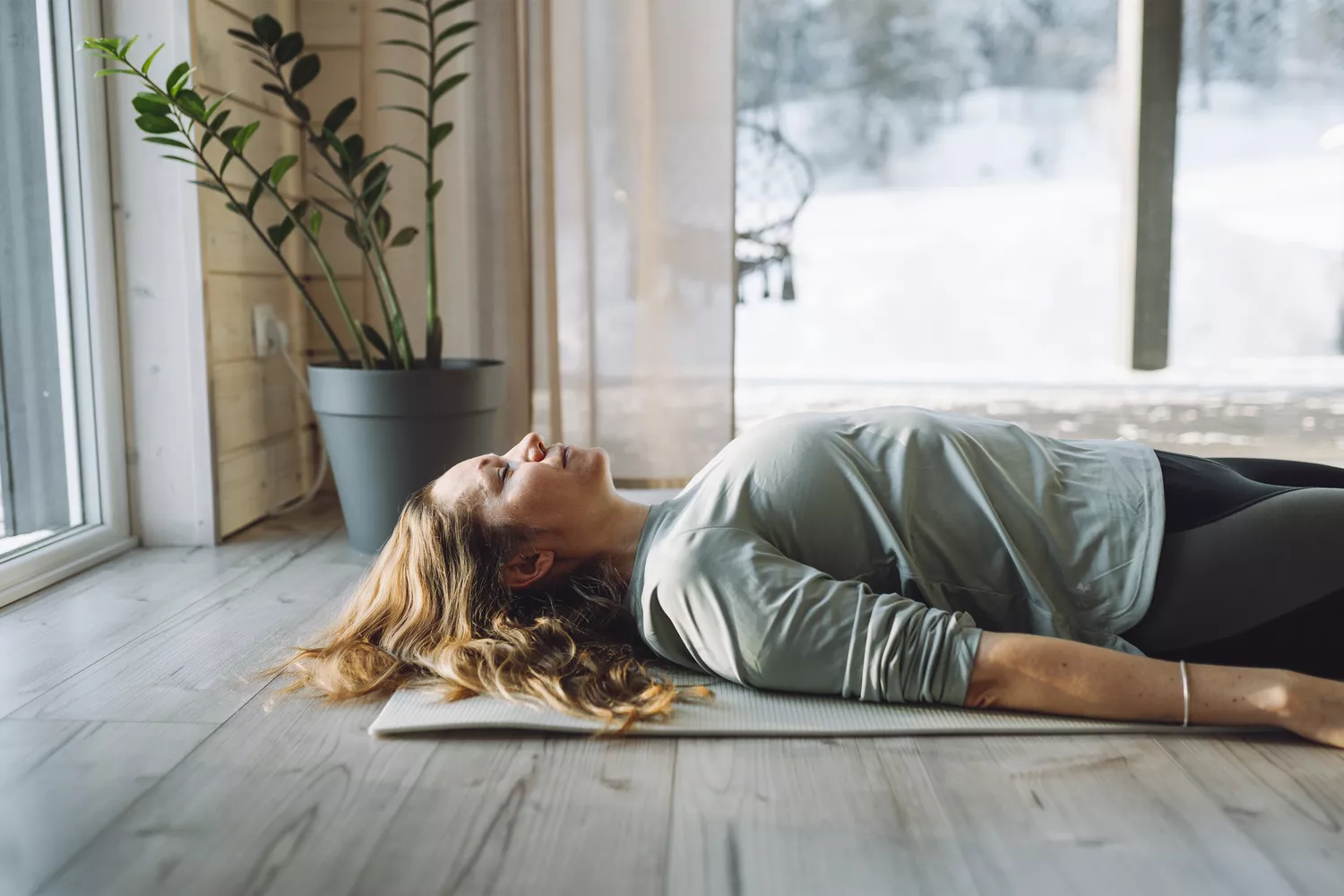 A woman lying in a relaxed Savasana pose on a yoga mat, eyes closed in a serene, bright room.
