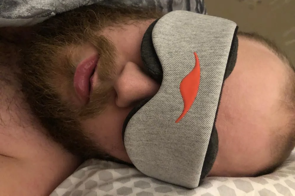 A man sleeping on his side while wearing a dark gray blackout eye mask.
