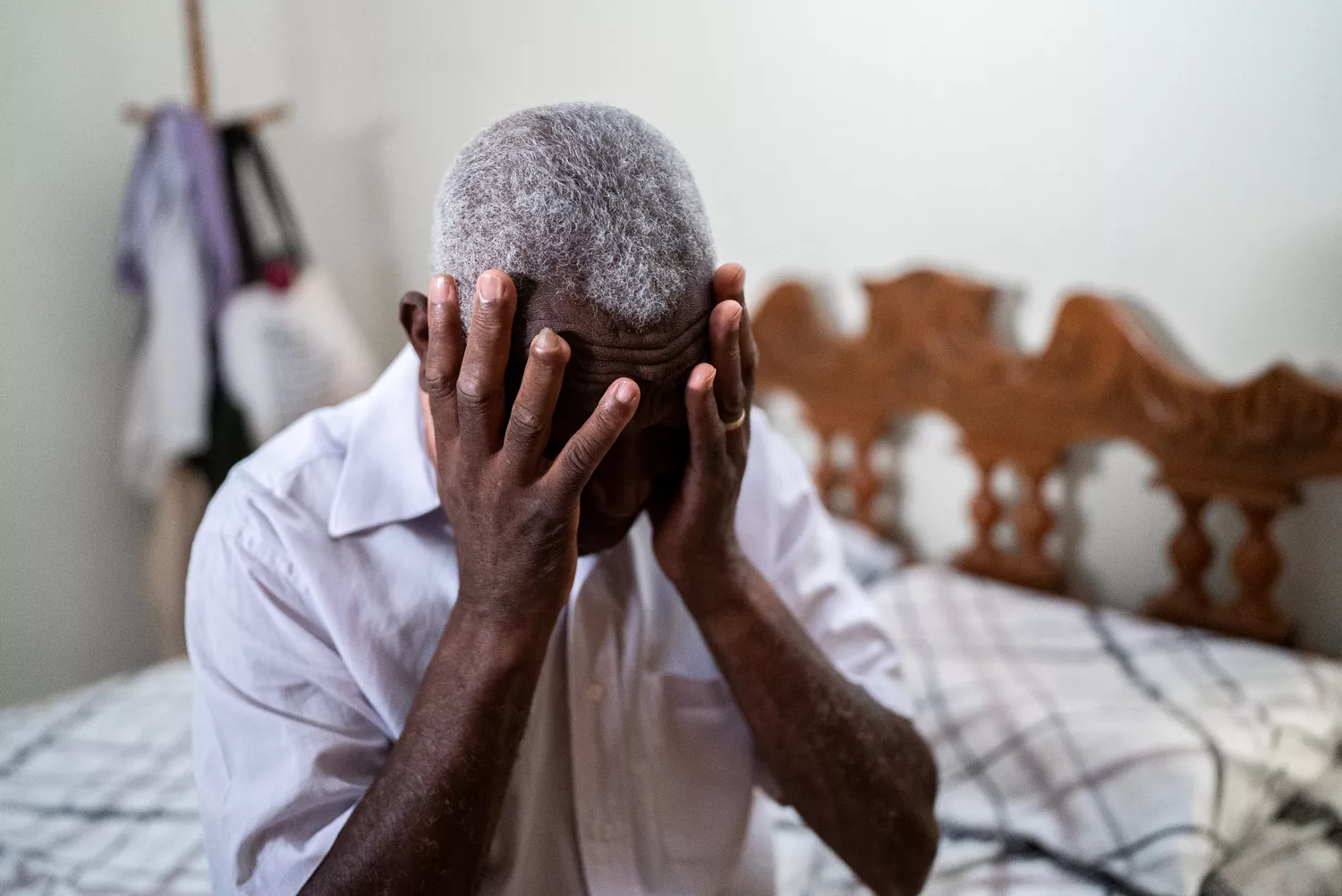 A senior man sitting on a bed with his hands covering his eyes and forehead, suggesting contemplation of health decisions.