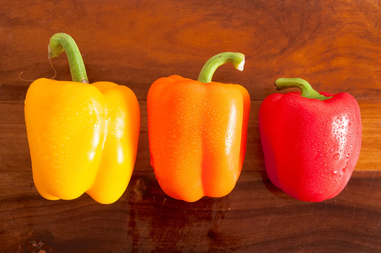 Bright red, yellow, and orange bell peppers on a wooden board.