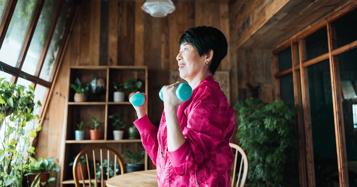 A smiling senior woman performing bicep curls with blue dumbbells in a home environment.