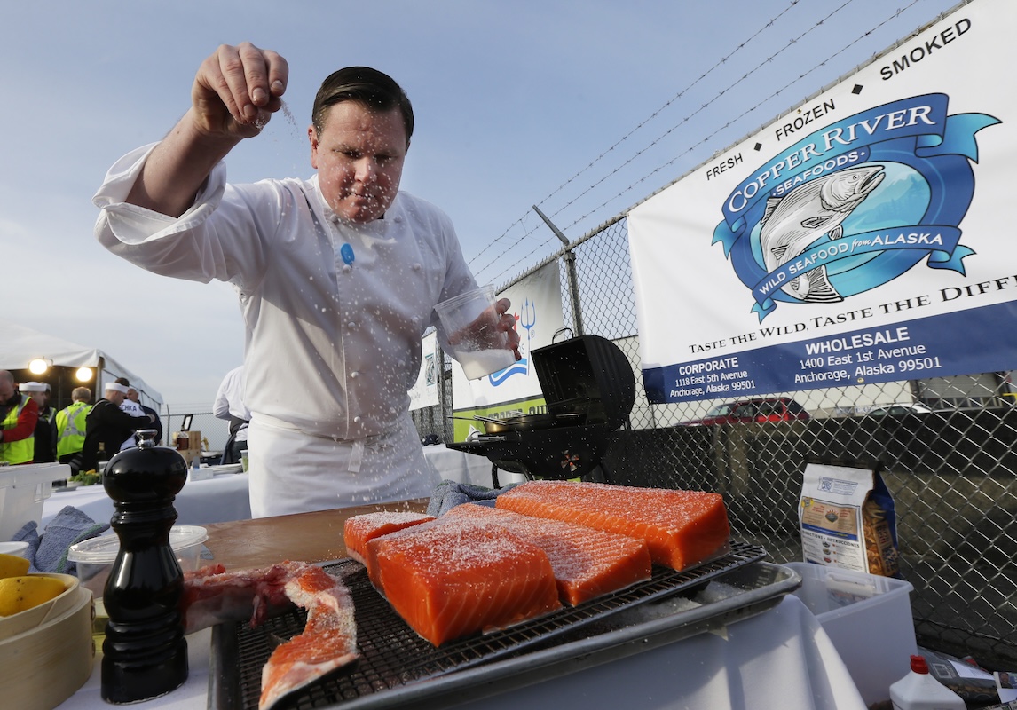 A professional chef sprinkling seasoning onto a fresh fillet of salmon.